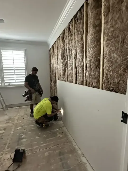 Two workers remove wet wall materials beside exposed framing and insulation inside a room