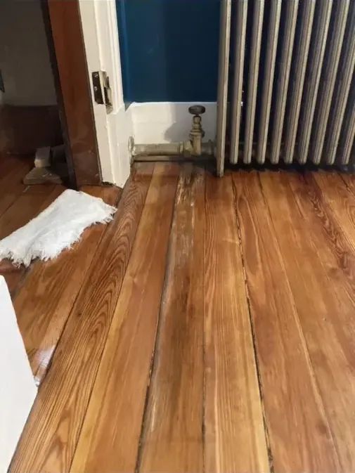 Damp wood flooring beneath a radiator beside an open wall and loose baseboard