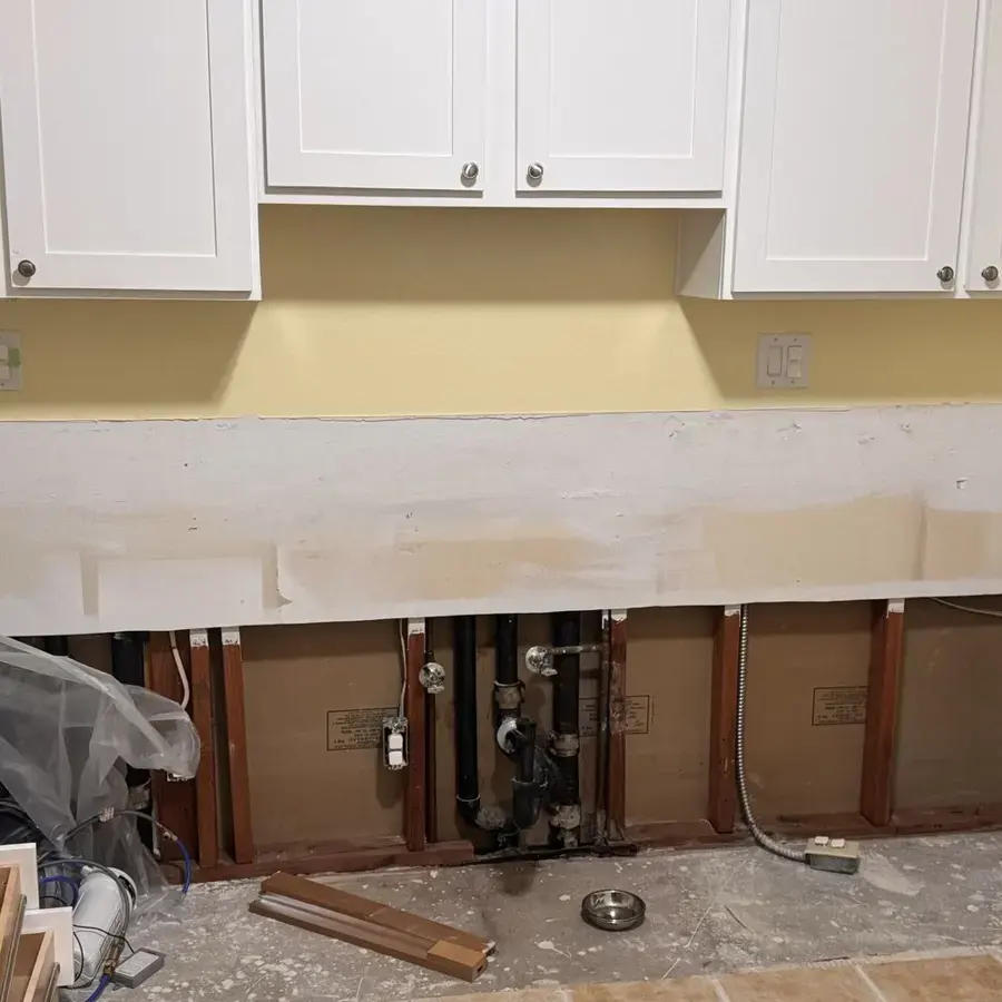 Open wall framing and exposed pipes beneath white upper cabinets in a kitchen
