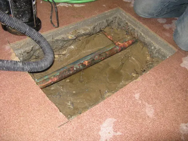Copper water pipe exposed in a trench cut through a concrete floor, with a vacuum hose at the opening