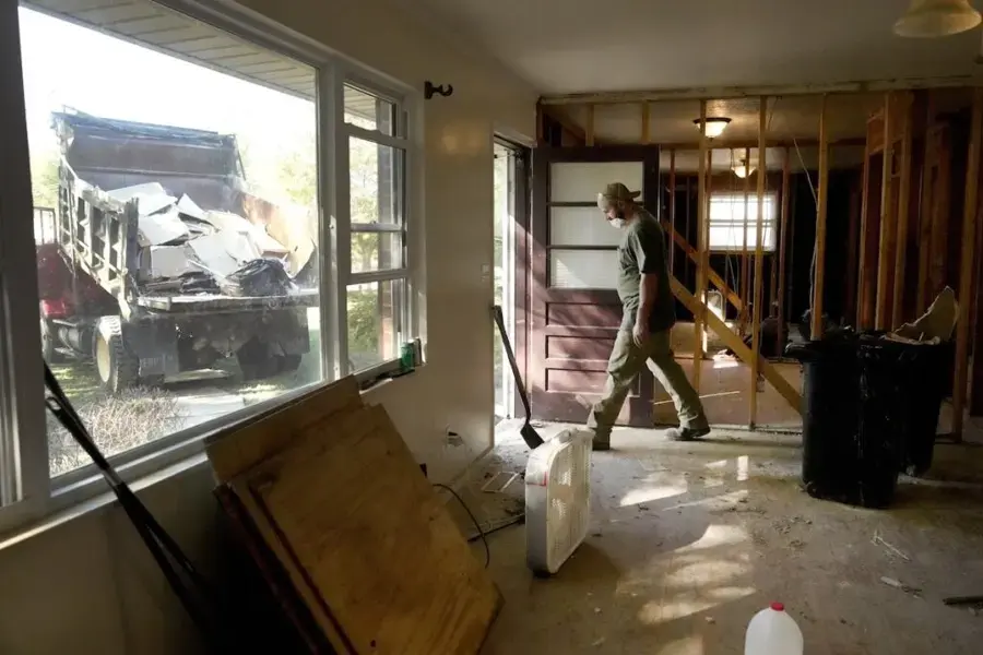 A worker removes damaged interior materials beside exposed wall framing inside a house